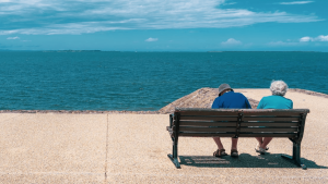 Two elderly people sit on a bench facing a calm blue sea along a seawall.