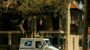 USPS delivery van on a tree-lined street in front of a beige, columned building with a balcony; a hanging American flag is visible behind the tree.