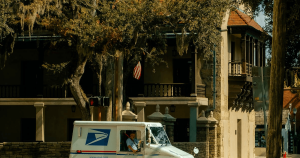 White delivery van drives past a beige, columned building under a large tree; a red-and-white striped flag hangs nearby.